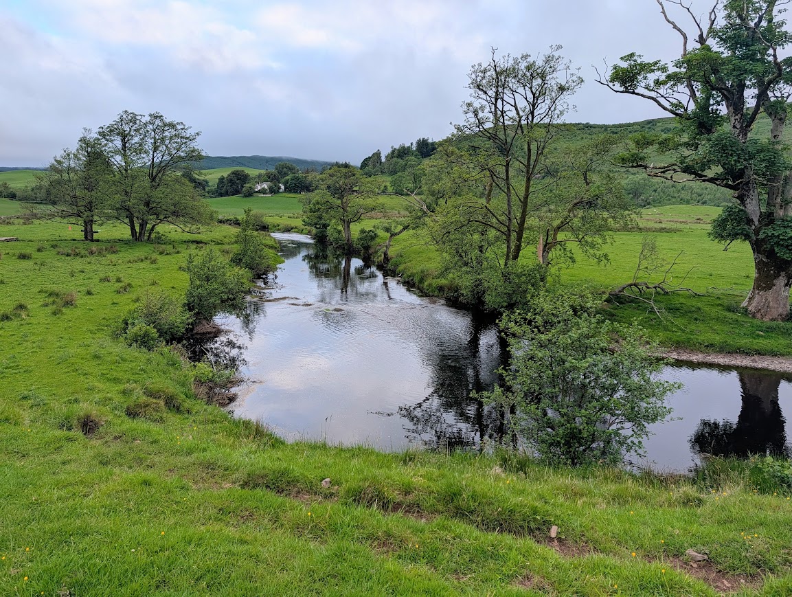The Water of Girvan reflecting the sky on a summer evening, surrounded by lush green fields. 