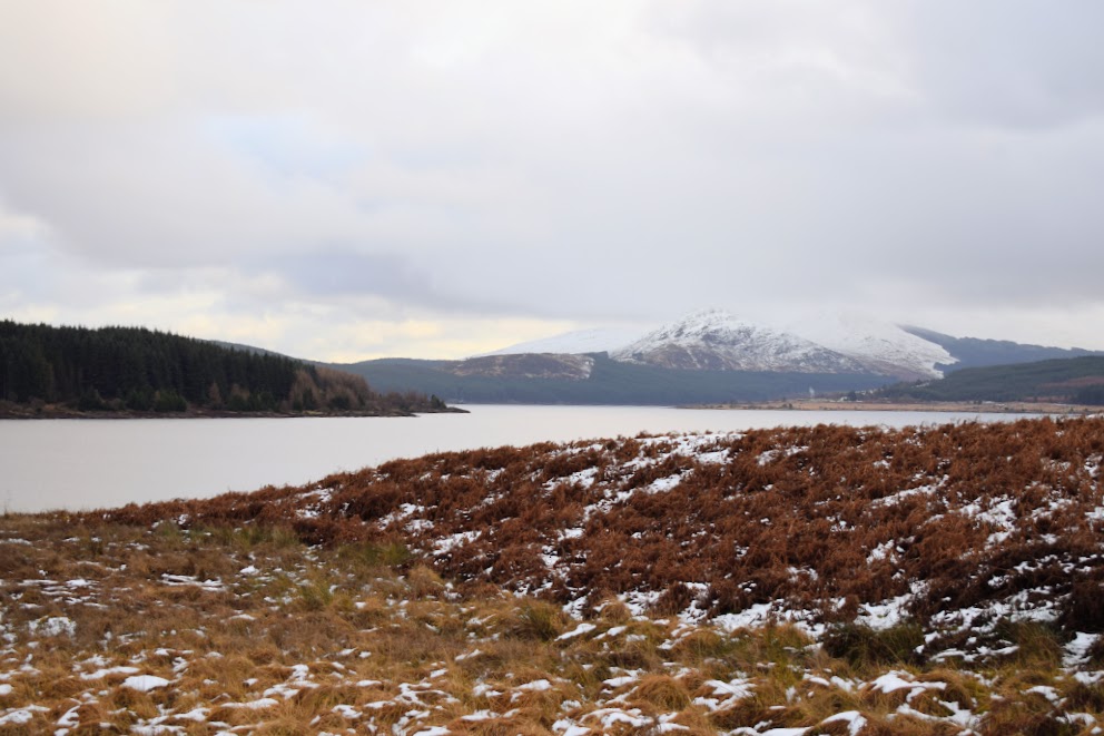 Patchy snow covers moorland scrub at the side of a large expanse of water: Loch Doon. The surrounding rolling hills are snow-capped and shrouded in low cloud