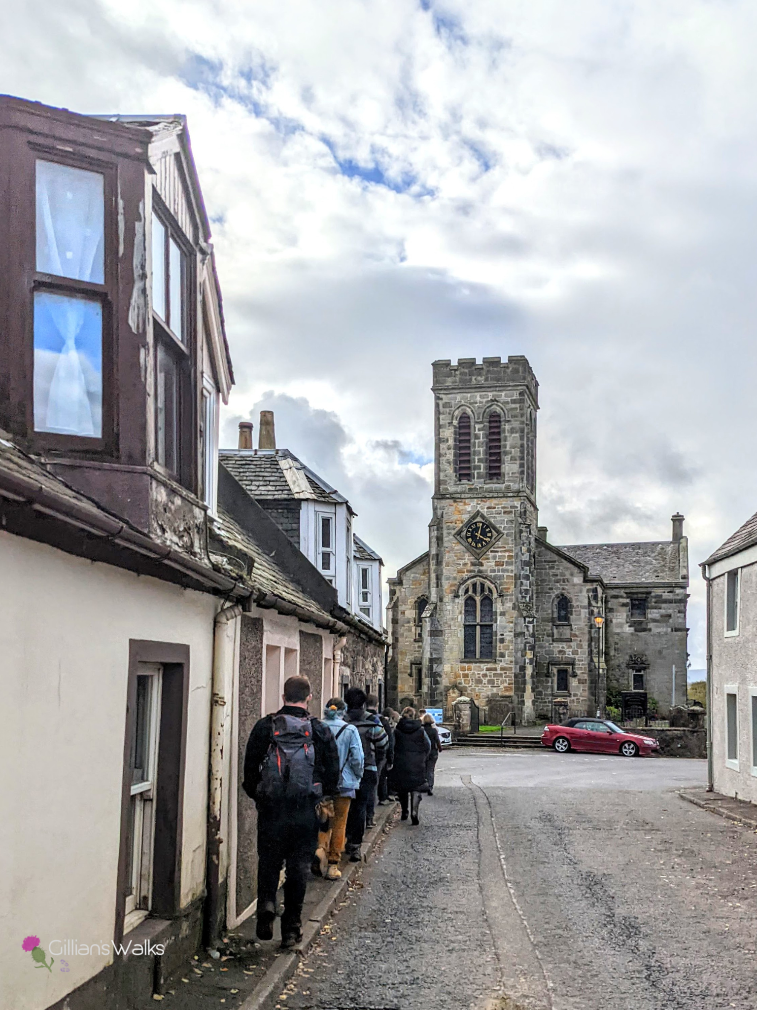 A group of people walking single file along the narrow pavement on Main Street in Dunlop.