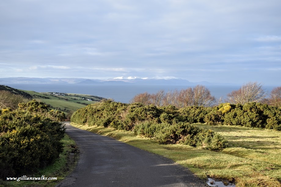 Single track road winding downhill towards a snow-capped Isle of Arran
