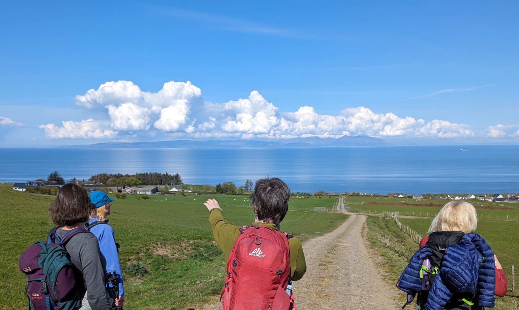 Four walkers stop on the hillside to admire the view. A female walker points out to sea. The water is flat-calm and the sky blue, with a cluster of white fluffy clouds over the Isle of Arran. 