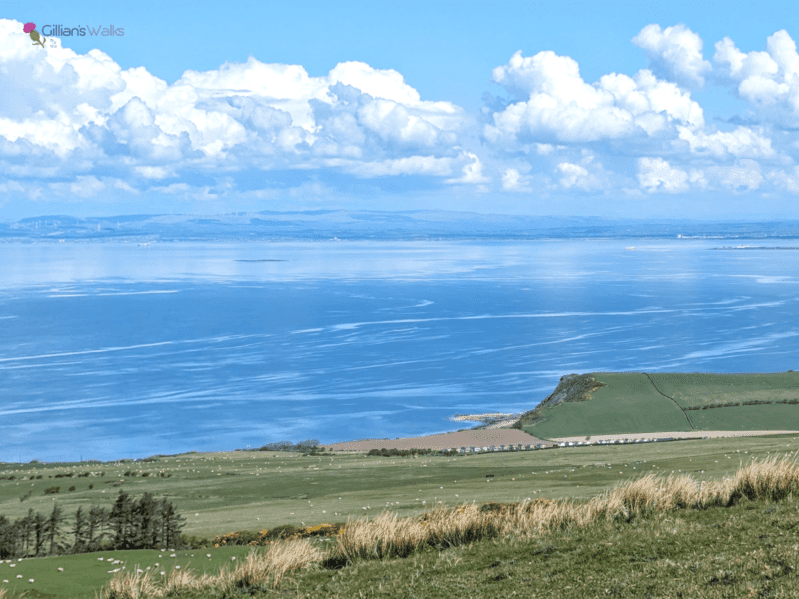View from summit of Brown Carrick Hill on a calm, clear day - lush farmland in the foreground, leading down to the dramatic Heads of Ayr cliffs and the glass-like water of the firth of clyde. 