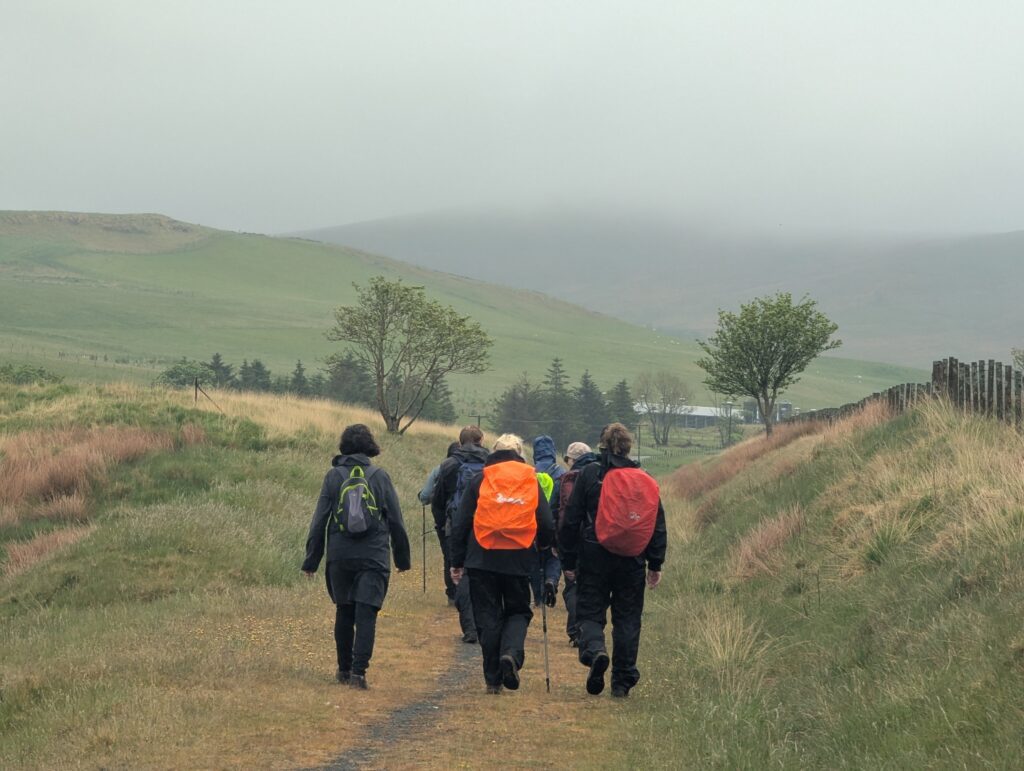 Group of hikers walking along a grassy track on a drizzly day, low cloud covering the top of the hills ahead.