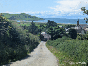 A concrete single track road winds downhill to a cluster of buildings, beyond which the sea is visible and the distant grassy slopes of Downan Hill