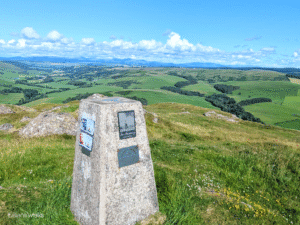 Stone trig point at the top of Knockdolian Hill, with memorial plaques attached to the two visible sides