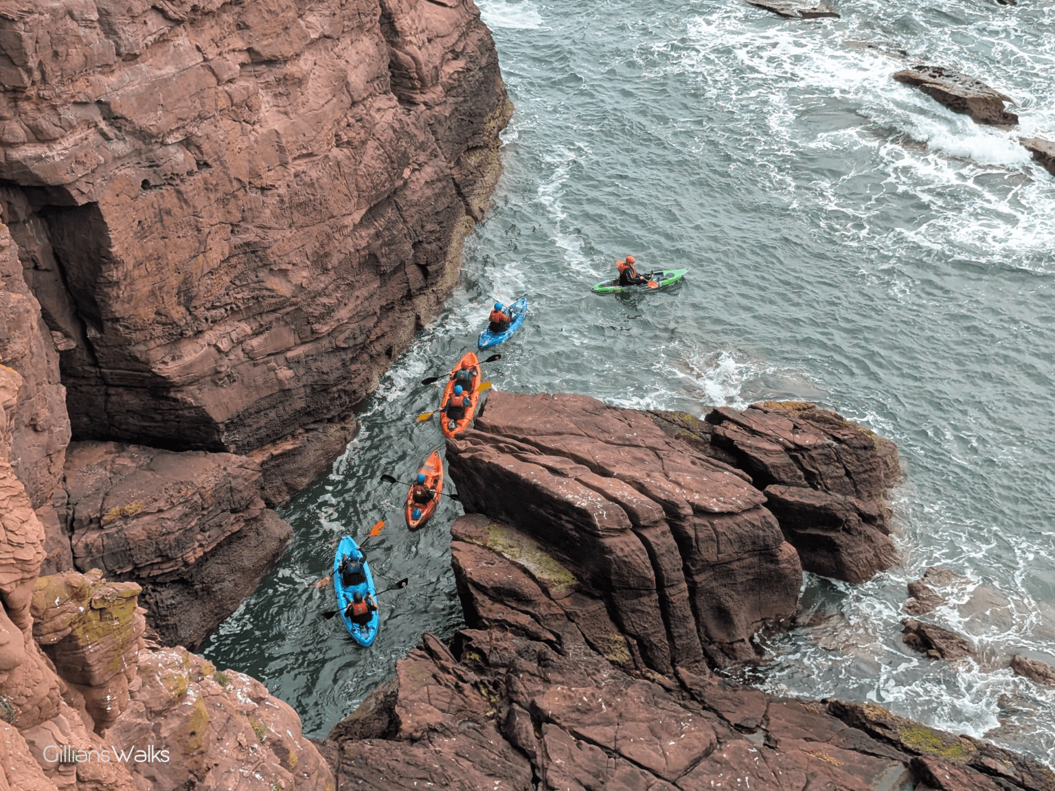 Five people on kayaks in the sea directly below me, entering a narrow channel between the cliffs