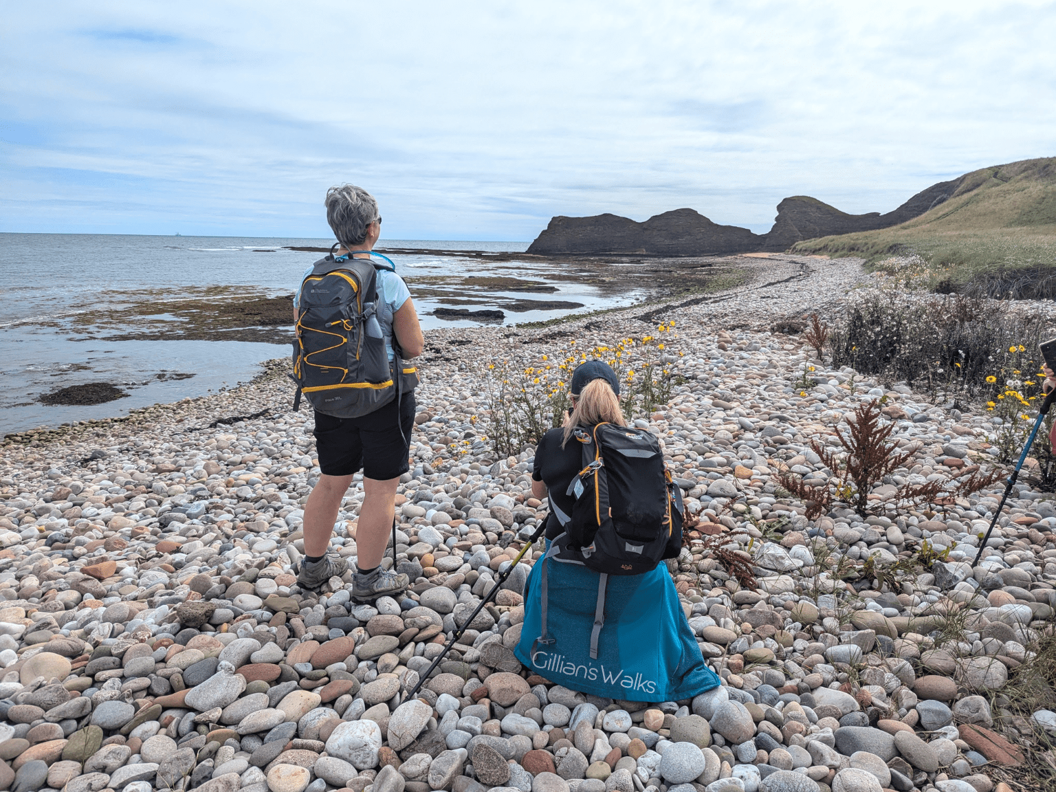 Two walkers pausing to admire the view on the pebble beach at Carlingheugh Bay, one of them crouching down to take a photo