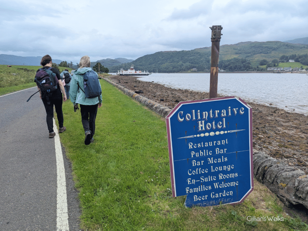 walkers on the side of a road with a metal sign at the bottom of a post advertising the Colintraive Hotel