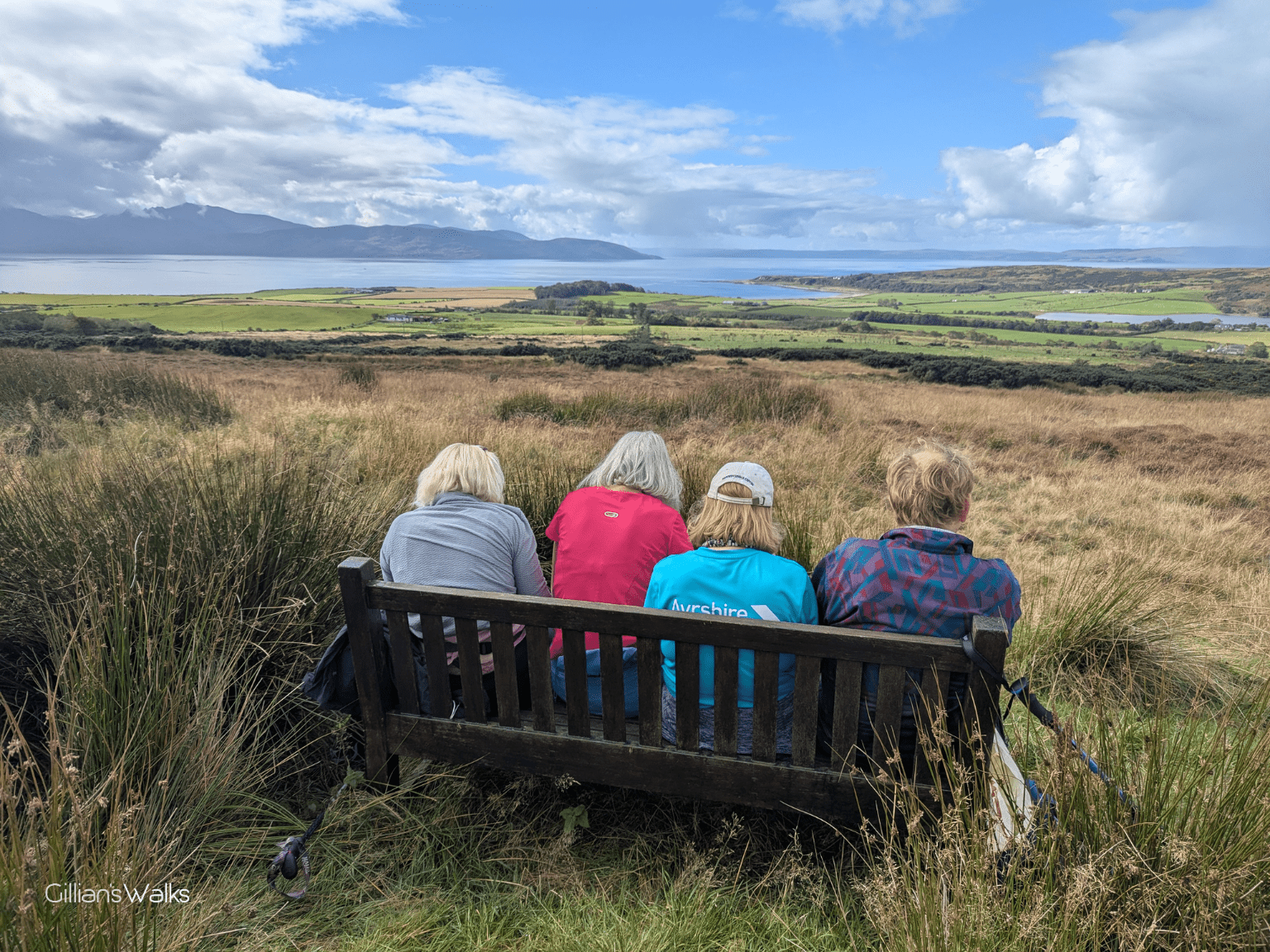 Four walkers squashed onto a small bench overlooking moorland, farmland, the coast and part of Arran