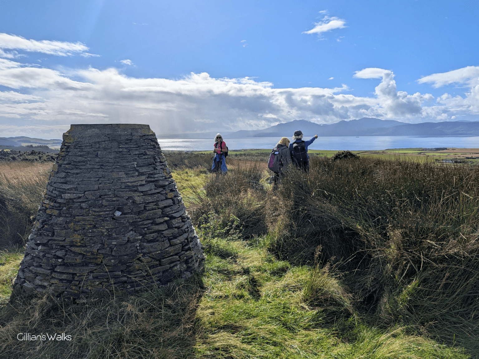 A stone cairn in the foreground, and the silhouette of two people pointing towards the view over the sea to Arran