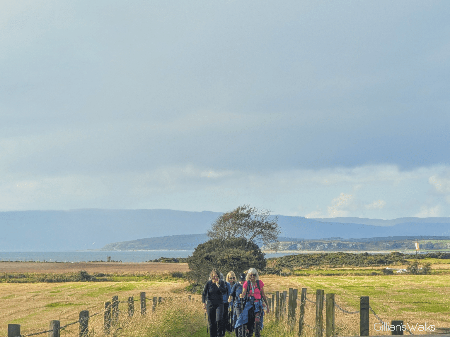 A group of walkers coming towards the camera, a backdrop of farmland and rugged coastline