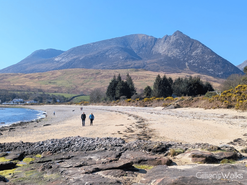 A swathe of sandy beach with a backdrop of high mountains against blue sky. Two people are walking on the beach.
