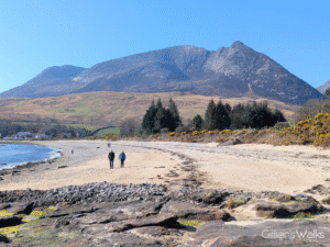 A swathe of sandy beach with a backdrop of high mountains against blue sky. Two people are walking on the beach.