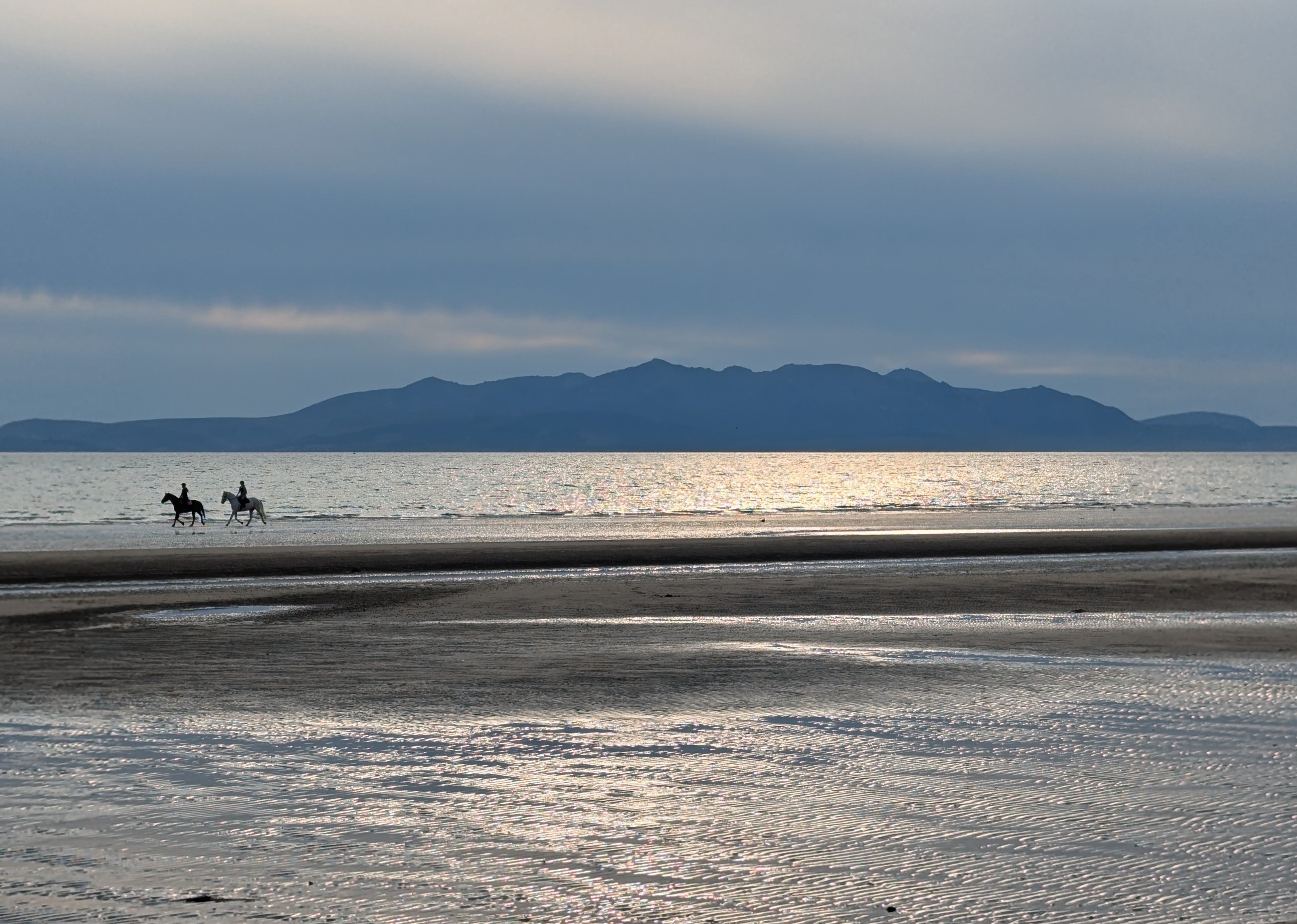 Silhouette of the Arran mountains against the shimmering sea and wet sandy beach. Two horseriders are on the sand.