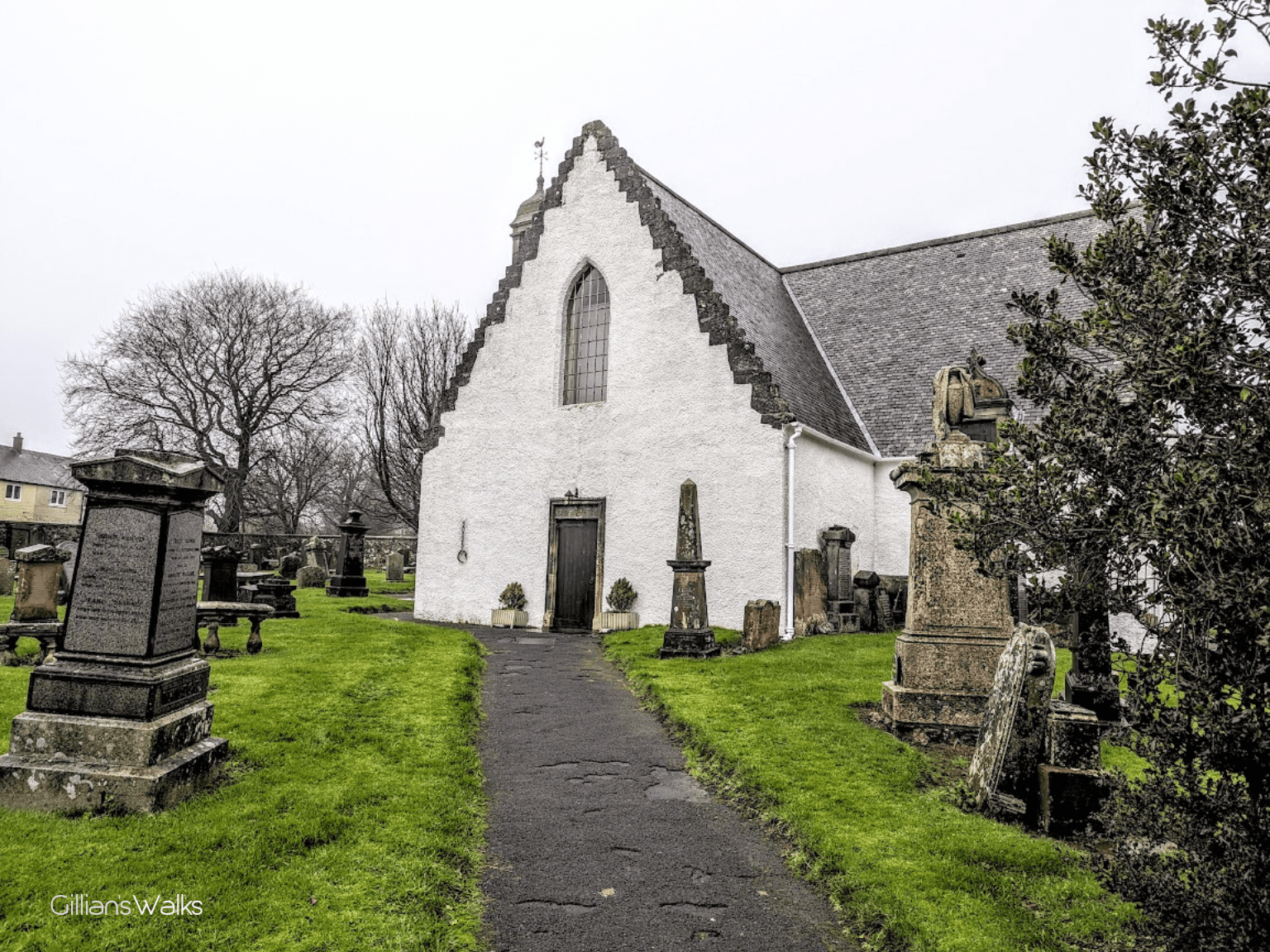 Historic white-rendered church with steep crow-stepped gable roof surrounded by an old graveyard featuring weathered stone headstones, obelisks, and moss-covered monuments. A narrow black path leads to a dark wooden door framed by two planters. Bare winter trees and green grass create a contrast against the overcast sky.
