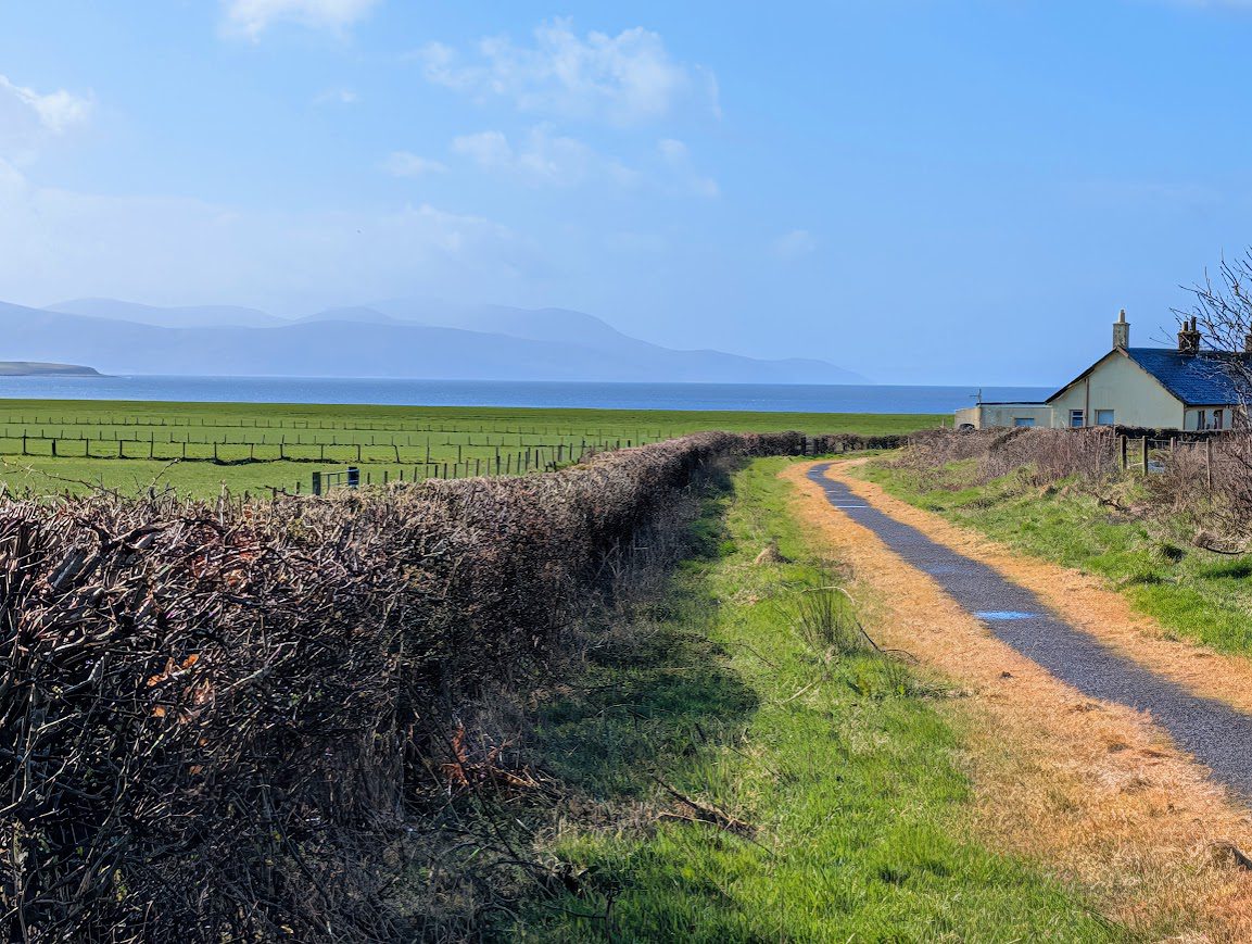 A narrow surfaced path with open views across lush farmland towards a silhouette of the Isle of Arran.