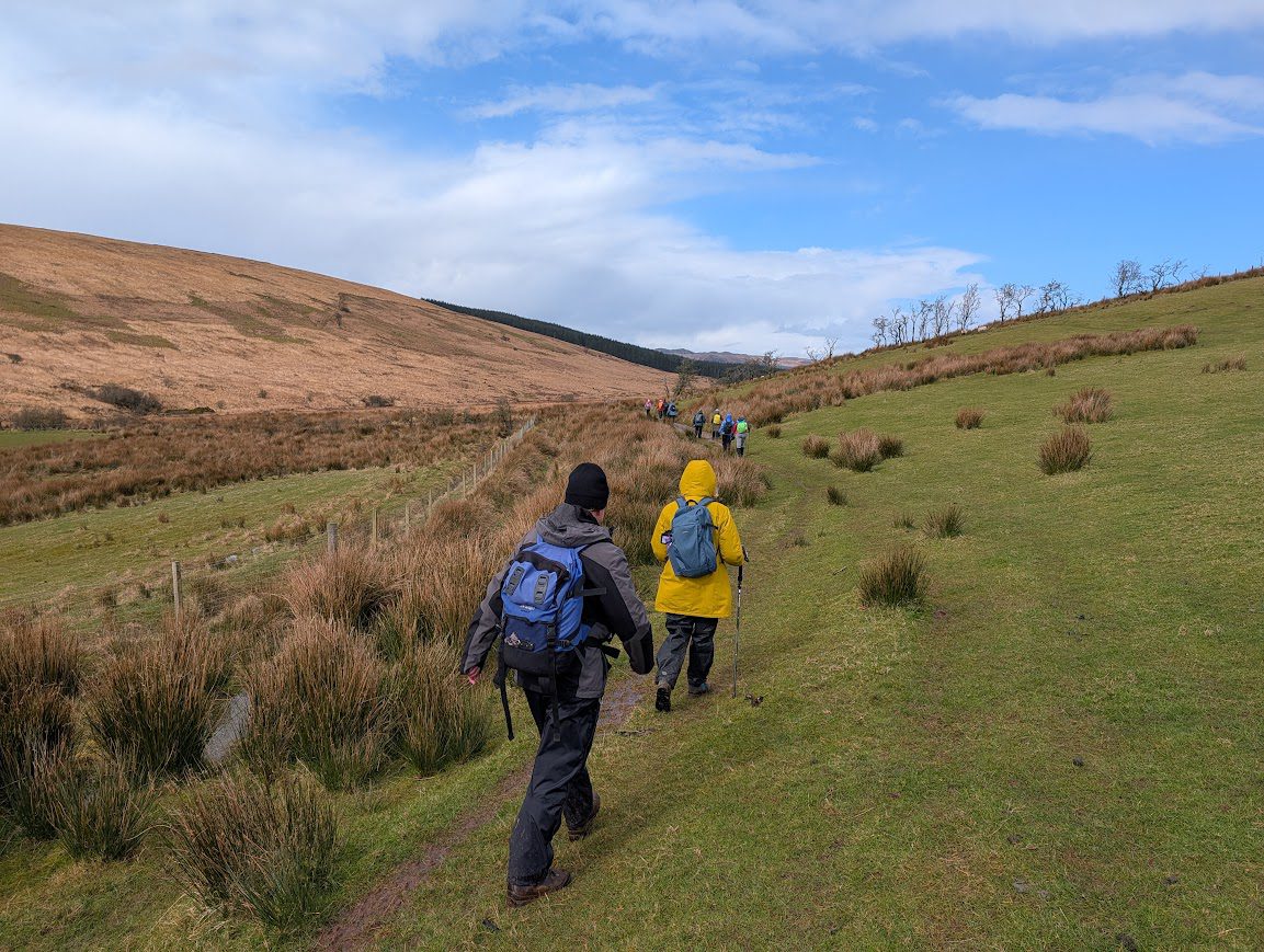 A group of walkers crossing a field on short grass with the occasional tussocks indicating marshy ground