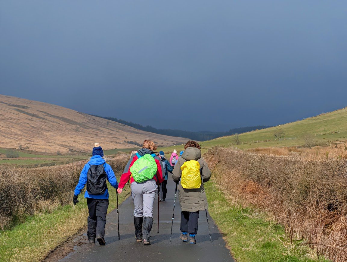 A group of walkers on a country road with the sun on their backs, but heading towards a looming dark sky