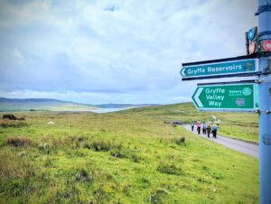 Metal pole with metal signage pointing to the left, with the words 'Gryffe Reservoirs' on the top sign, and 'Gryffe Valley Way, Rotary' on the bottom. In the background is open grassy moorland and a loch, with a group of walkers approaching along a track