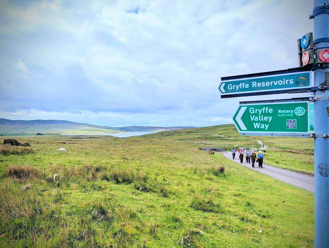 Metal pole with metal signage pointing to the left, with the words 'Gryffe Reservoirs' on the top sign, and 'Gryffe Valley Way, Rotary' on the bottom. In the background is open grassy moorland and a loch, with a group of walkers approaching along a track