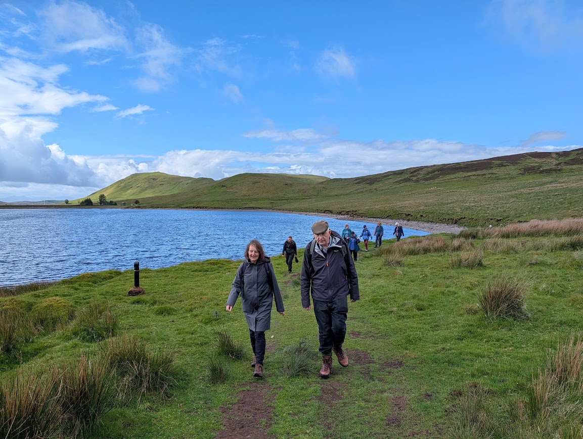 A group of walkers on a grassy track alongside a large loch backed by rolling green hills
