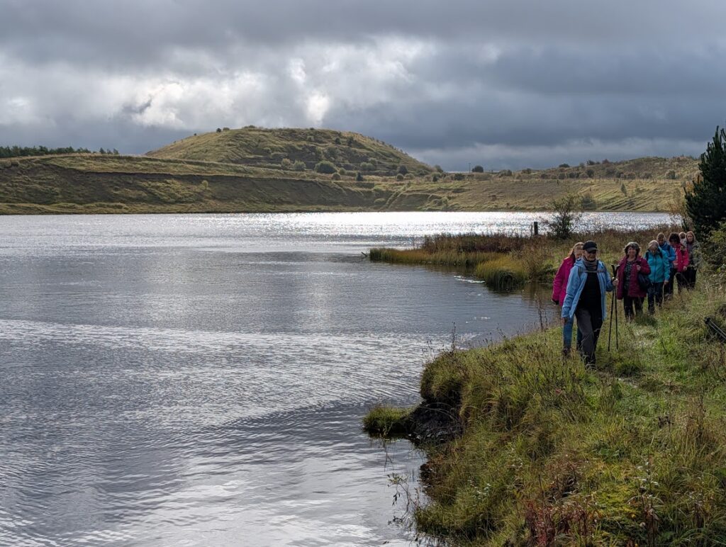 Several ladies walking along the banks of the loch, with one of the old coal bings in the backgroundf