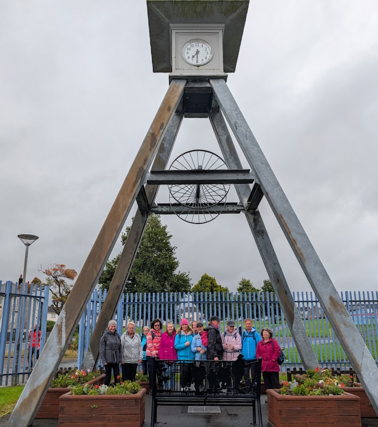 Group of walkers pose beneath a large steel A-frame from the coal mining era, with an analogue clock at the top.